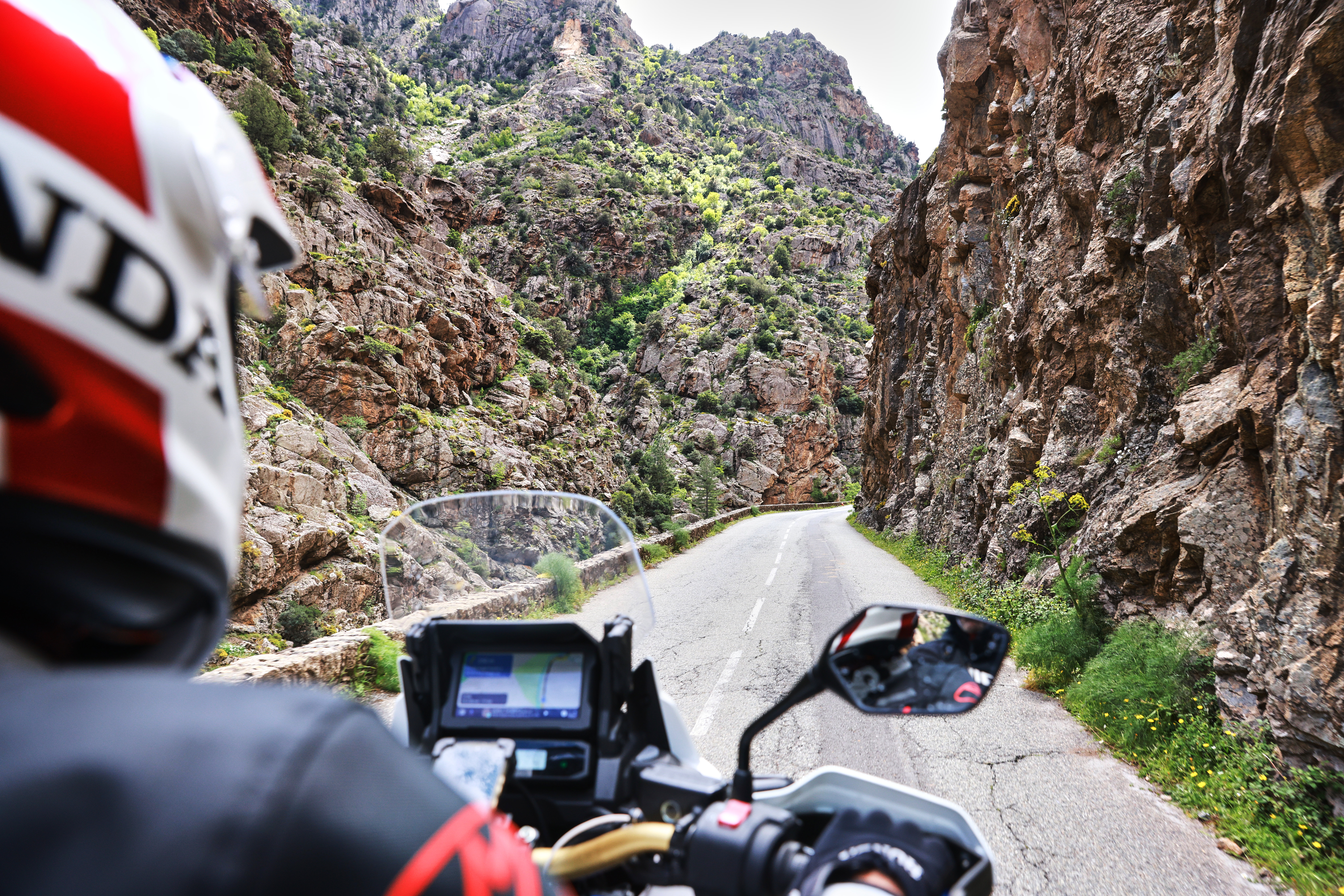 Vista dalla parte posteriore di una moto che percorre una strada stretta circondata da ripide scogliere rocciose e vegetazione verde.